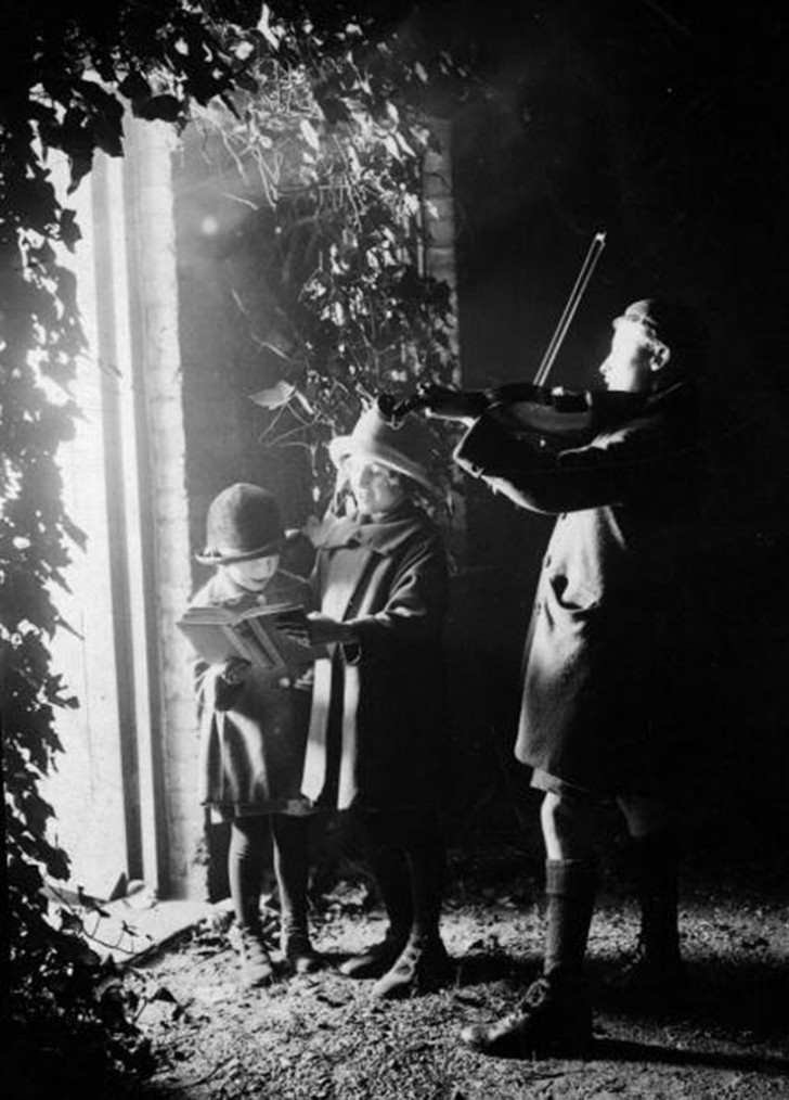 4. Tre bambini cantano cori di Natale accompagnati da un violino, davanti la porta di qualcuno, 1924.