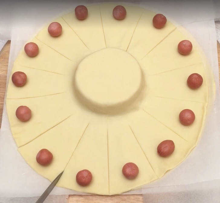 4. Tagliate degli spicchi di pasta senza arrivare al limite della forma di formaggio e adagiate un mini-würstel su ciascuno di essi.