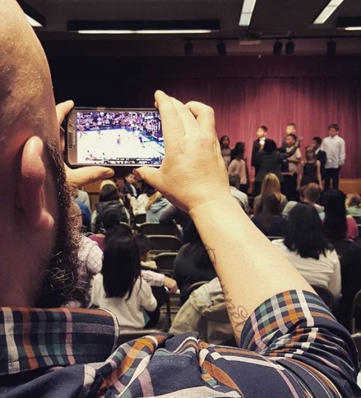 The man pretends to film his son at the school play, but instead is watching a basketball game!