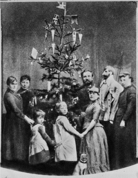 Una famiglia attorno all'albero di Natale, addobbato con bandiere e coni di carta contenenti noccioline e caramelle.
