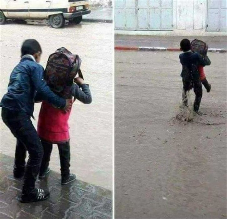 A boy picks up his younger brother to keep his shoes from getting wet since his brother has to spend all day at school.