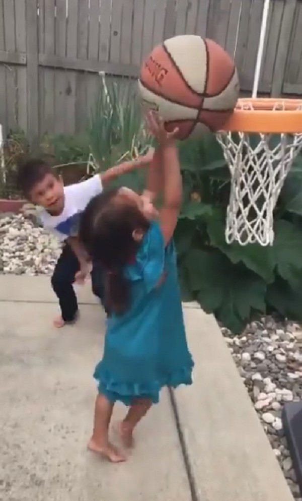 Deux frères et sœurs jouent au basket-ball dans le jardin : l'aîné surveille le tir de sa sœur.