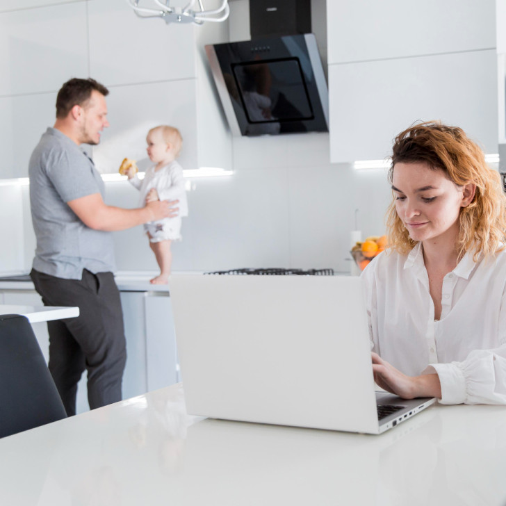 Donna al computer, sullo sfondo un papà con un neonato