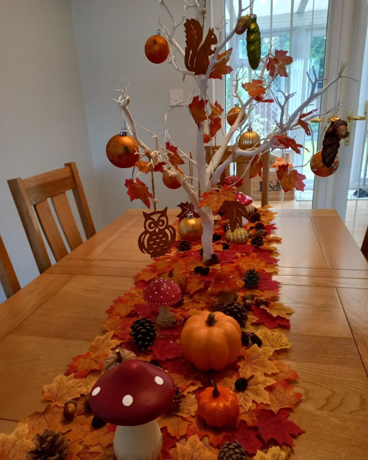 Een herfsttafeldecoratie met kleurrijke bladeren, paddenstoelen, pompoenen en een kale witte boom met oranje decoratieve ballen