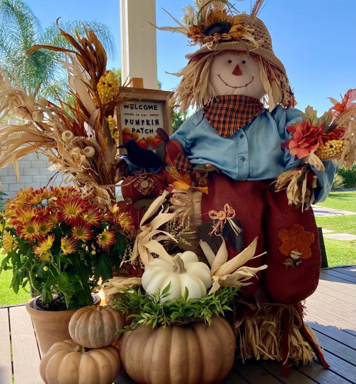 Composizione decorativa per il portico con spaventapasseri in paglia, spighe di grano e di mais, zucche e un grande vaso di fiori arancioni