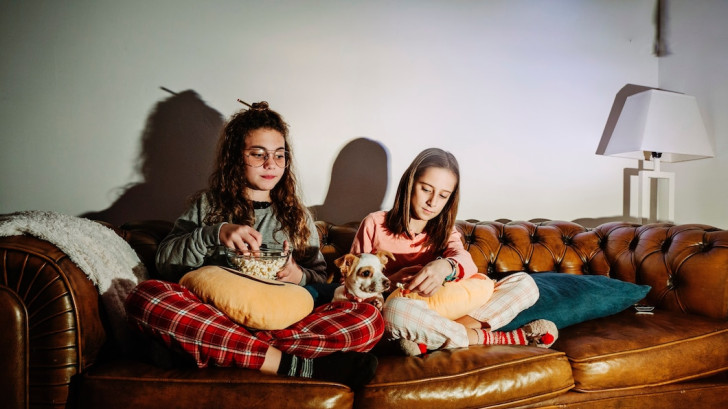 Due bambine su un divano, con snack, e il cane tra loro che guarda la TV