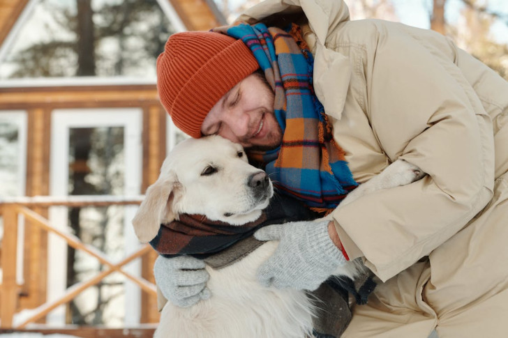 un uomo con indumenti invernali si china per abbracciare un cane a pelo bianco avvolto da una sciarpa