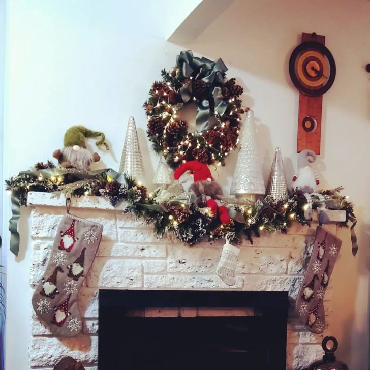 Felt gnomes set up next to silver pine trees, a wreath of pine cones, lights and decorated Christmas stockings