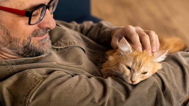 un uomo brizzolato sdraiato sul divano con in braccio un gatto rosso rilassato