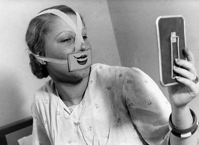 La didascalia della foto d'epoca: "Nella Scuola del Sorriso una donna guarda il suo volto allo specchio, recante una benda che le permette di sorridere".