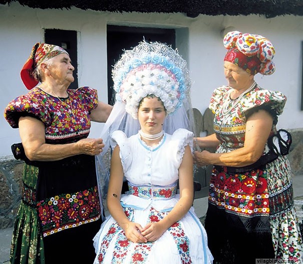 15. Een Hongaarse bruid draagt een jurk met bloemmotieven en tenminste drie levendige kleuren en verfijnde hoofddeksel met verweven korenaren maakt het compleet.