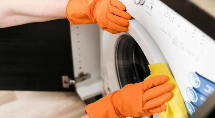 Person mit orangefarbenen Handschuhen beim Waschen der Außenseite der Waschmaschinentür