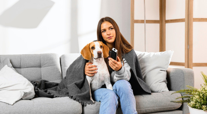 Una ragazza e un cane accomodati insieme sul divano con un plaid mentre guardano la TV