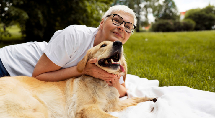 una donna con capelli bianchi che coccola un golden retriever, entrambi stesi su un telo in un parco