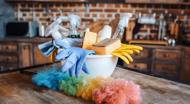 A kitchen table with a duster and a bowl containing sponges, cleaning products and rubber gloves