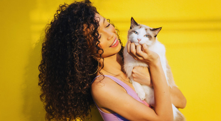 una donna con capelli lunghi, scuri e mossi tiene in braccio un gatto dal pelo chiaro e occhi azzurri che si lascia coccolare